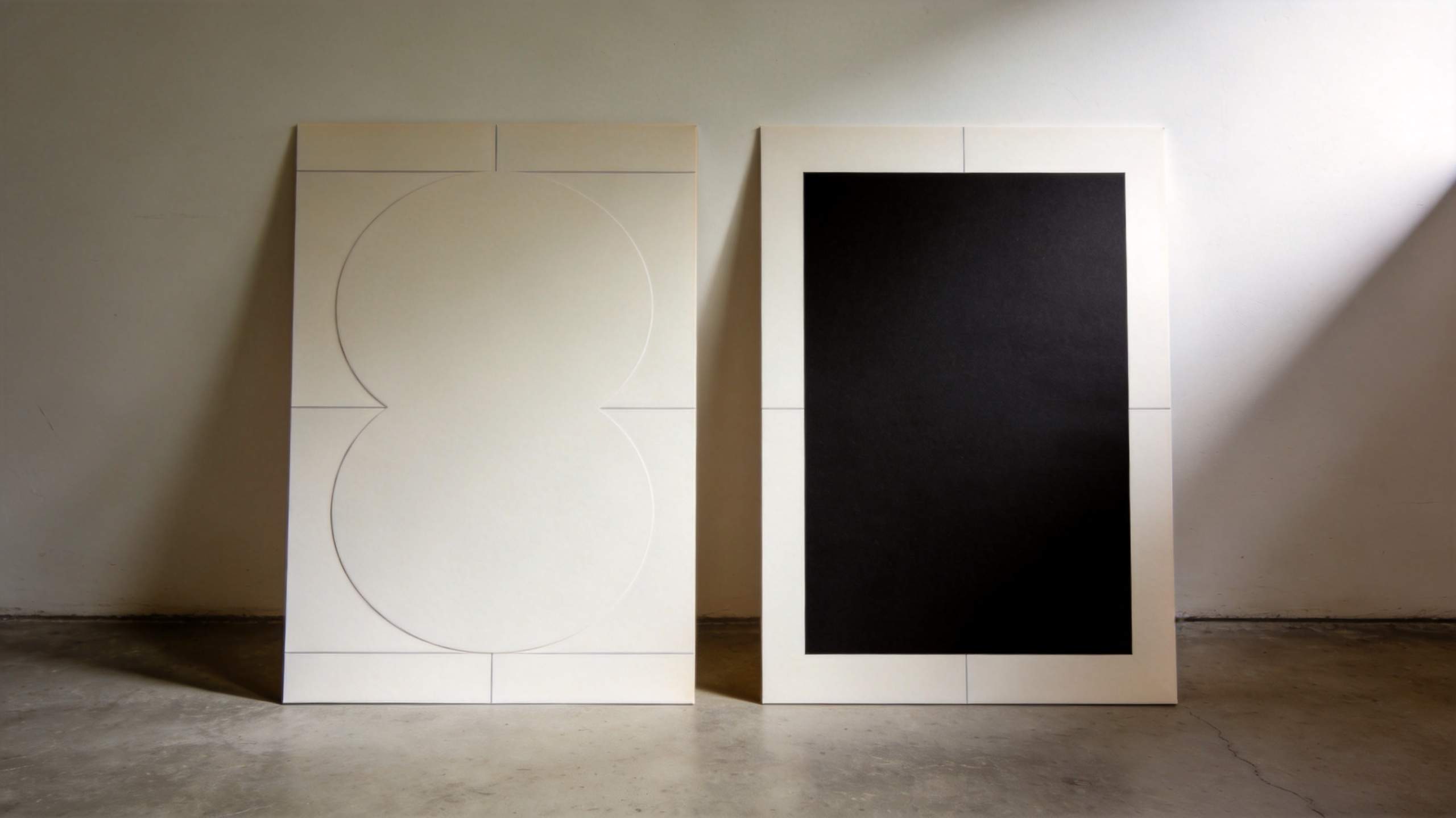 Two minimalist white panels lean against a wall, one with embossed circles and one with a large black rectangle.