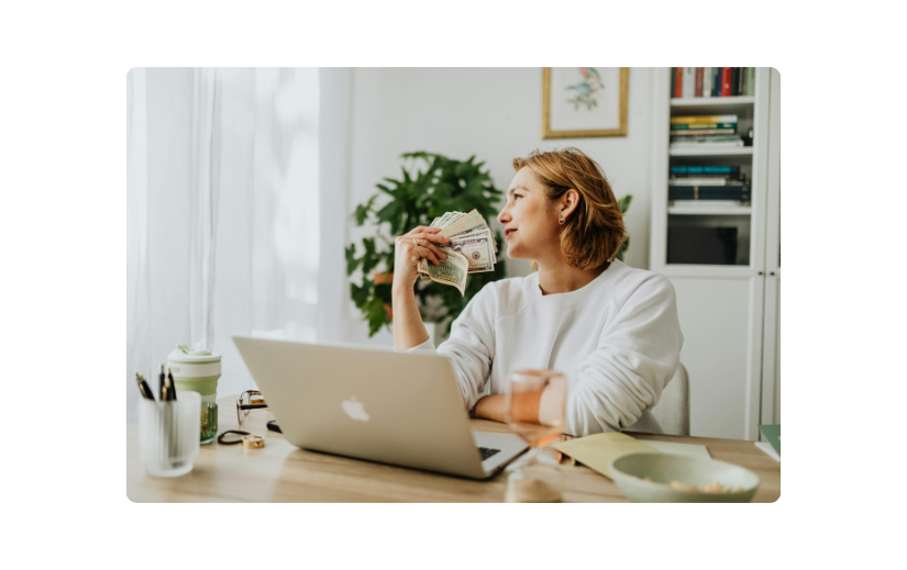 College student exploring side hustles while working on a laptop, holding cash in a home office setting. 