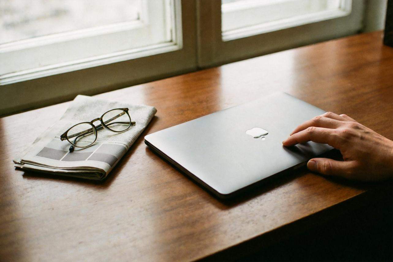 Hand using a laptop beside folded newspaper and eyeglasses on a wooden desk