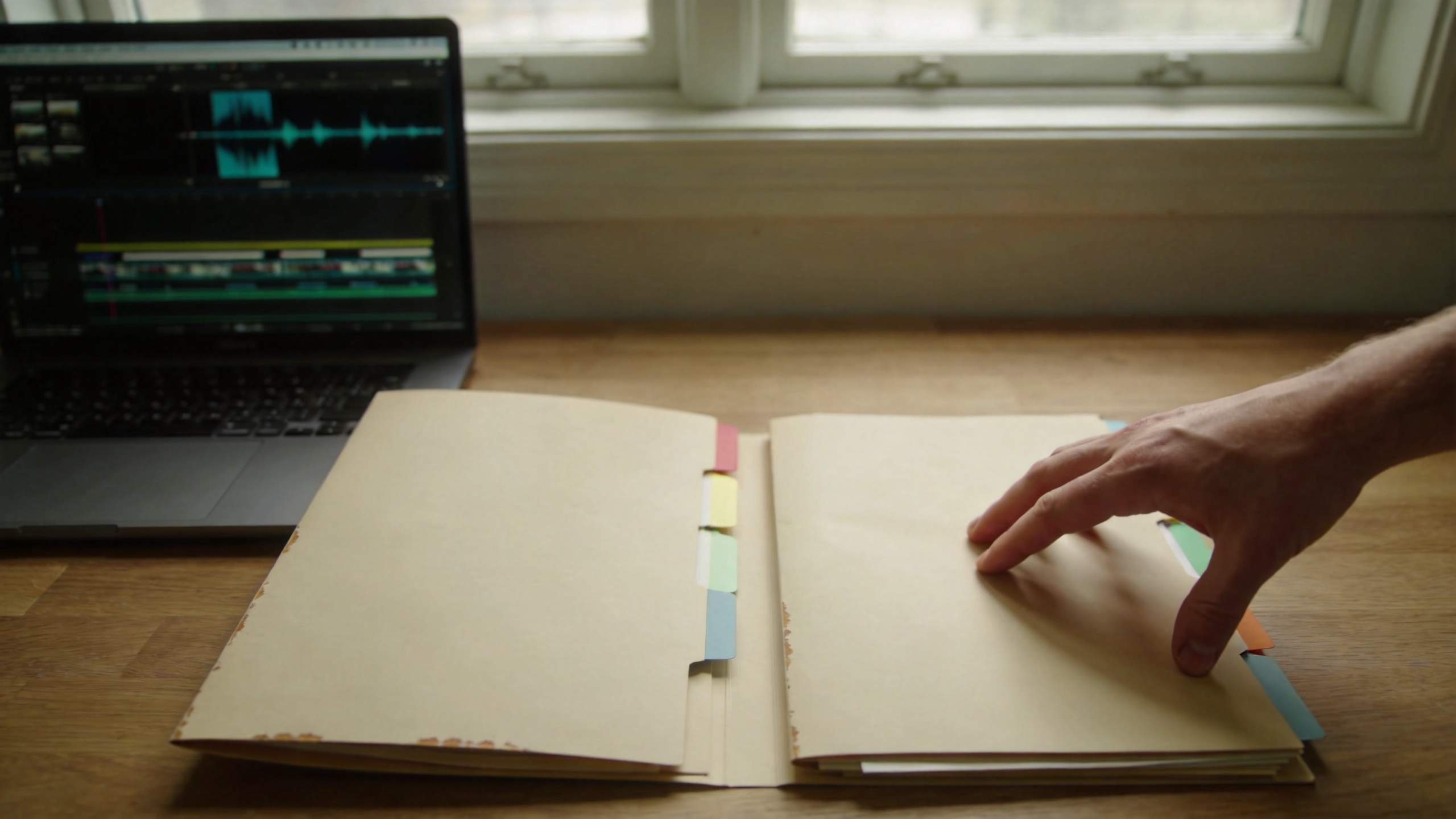 Hand turns page in a notebook beside a laptop with an audio editing timeline