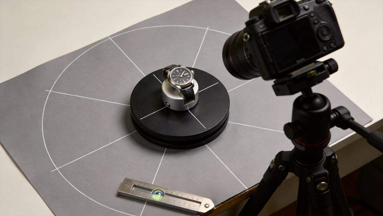 Camera on tripod filming a watch on a rotating turntable with a gray backdrop and measurement tools