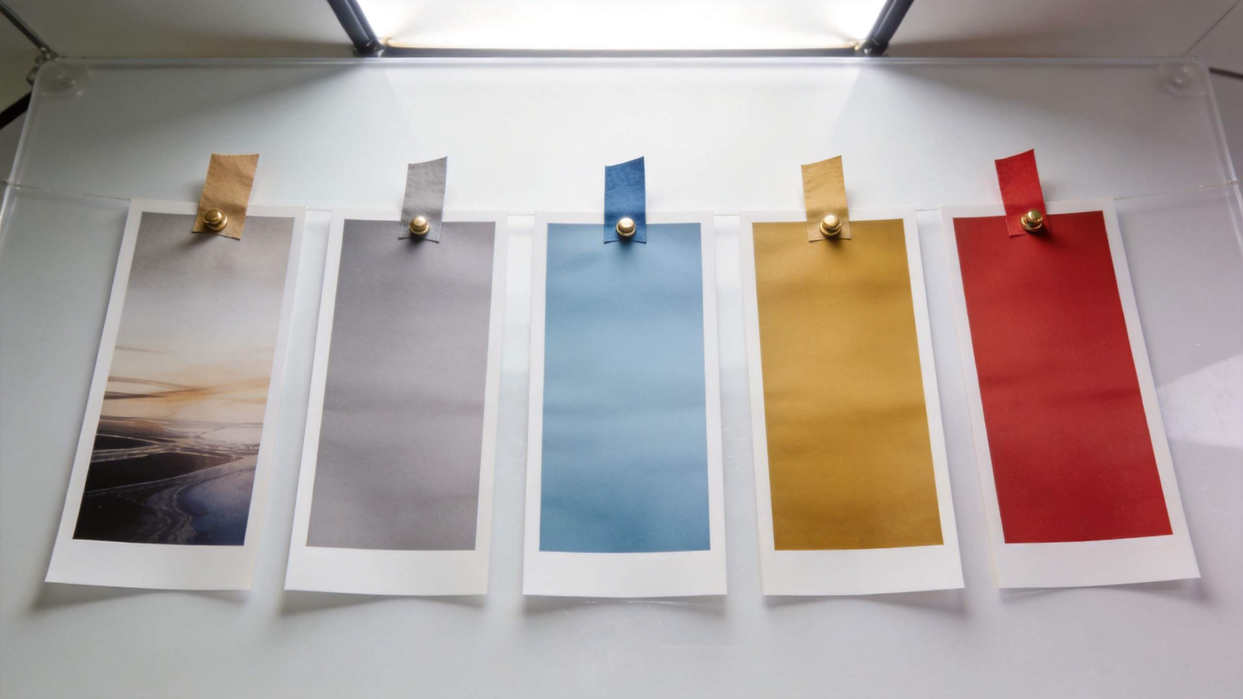 Five hanging polaroid-style prints clipped to a clear board in gray, blue, gold, and red