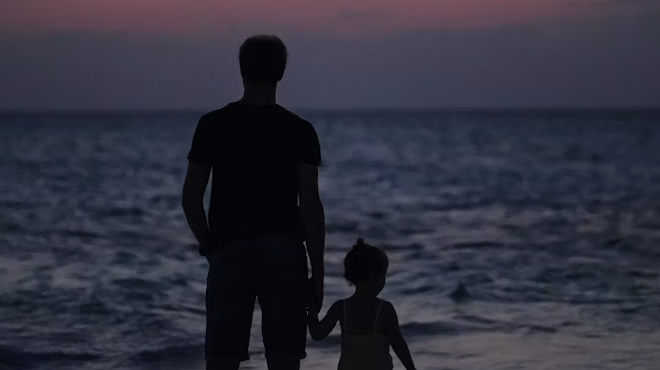A father and daughter walking hand-in-hand through a park at sunset.