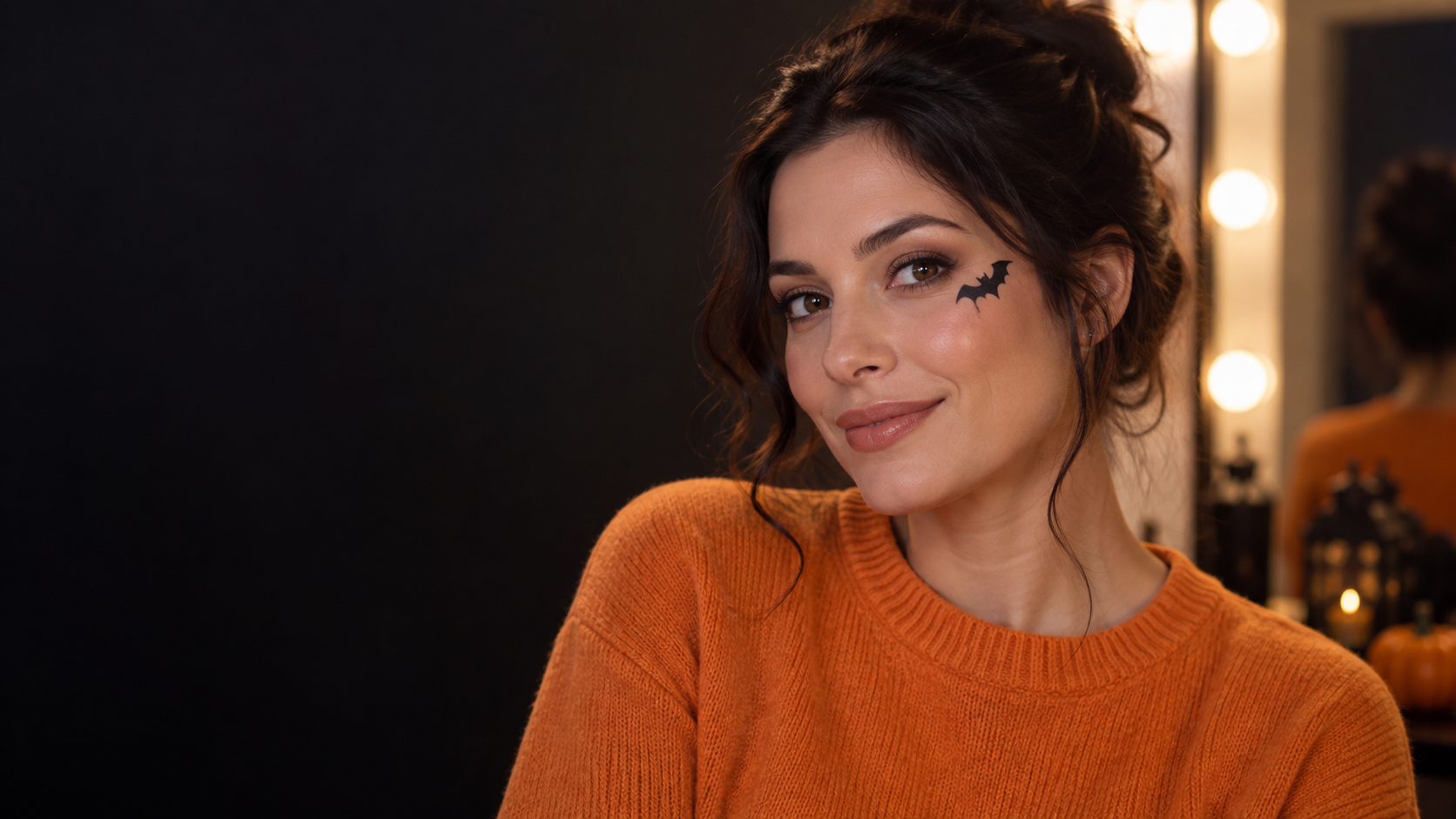 Tiny Bat Eyeliner: A fictional adult woman with a single small black bat wing drawn beside one eye, otherwise minimal makeup and a soft orange sweater, clean mirror-lit portrait.