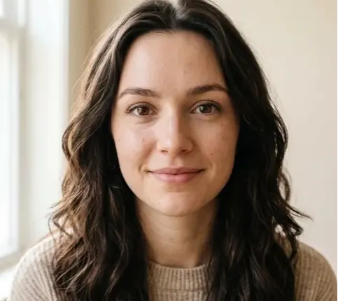Woman with dark wavy hair wearing a beige ribbed sweater against a light background