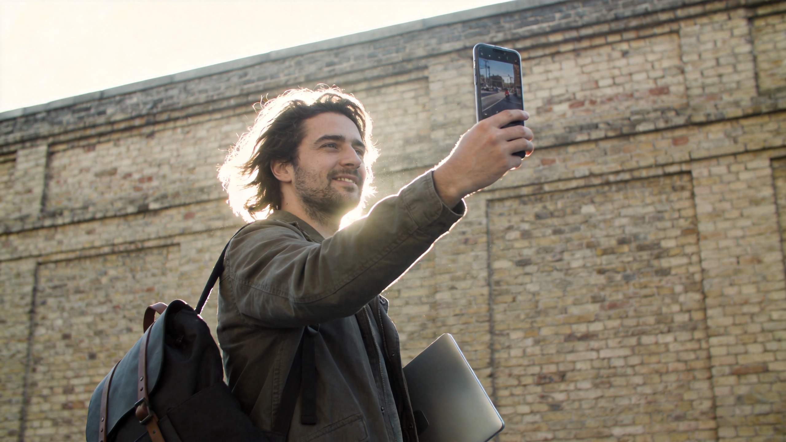 Man outdoors holding up a smartphone while carrying a laptop and backpack